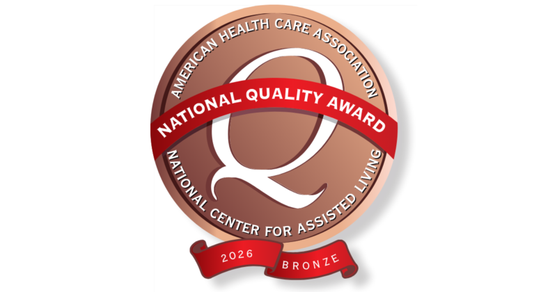 A bronze-colored circular badge with a white "Q" in the center. Text around the top reads "American Health Care Association," and around the bottom "National Center for Assisted Living." A red ribbon across the center says "National Quality Award." A smaller red banner at the bottom indicates "2026" and "Bronze."